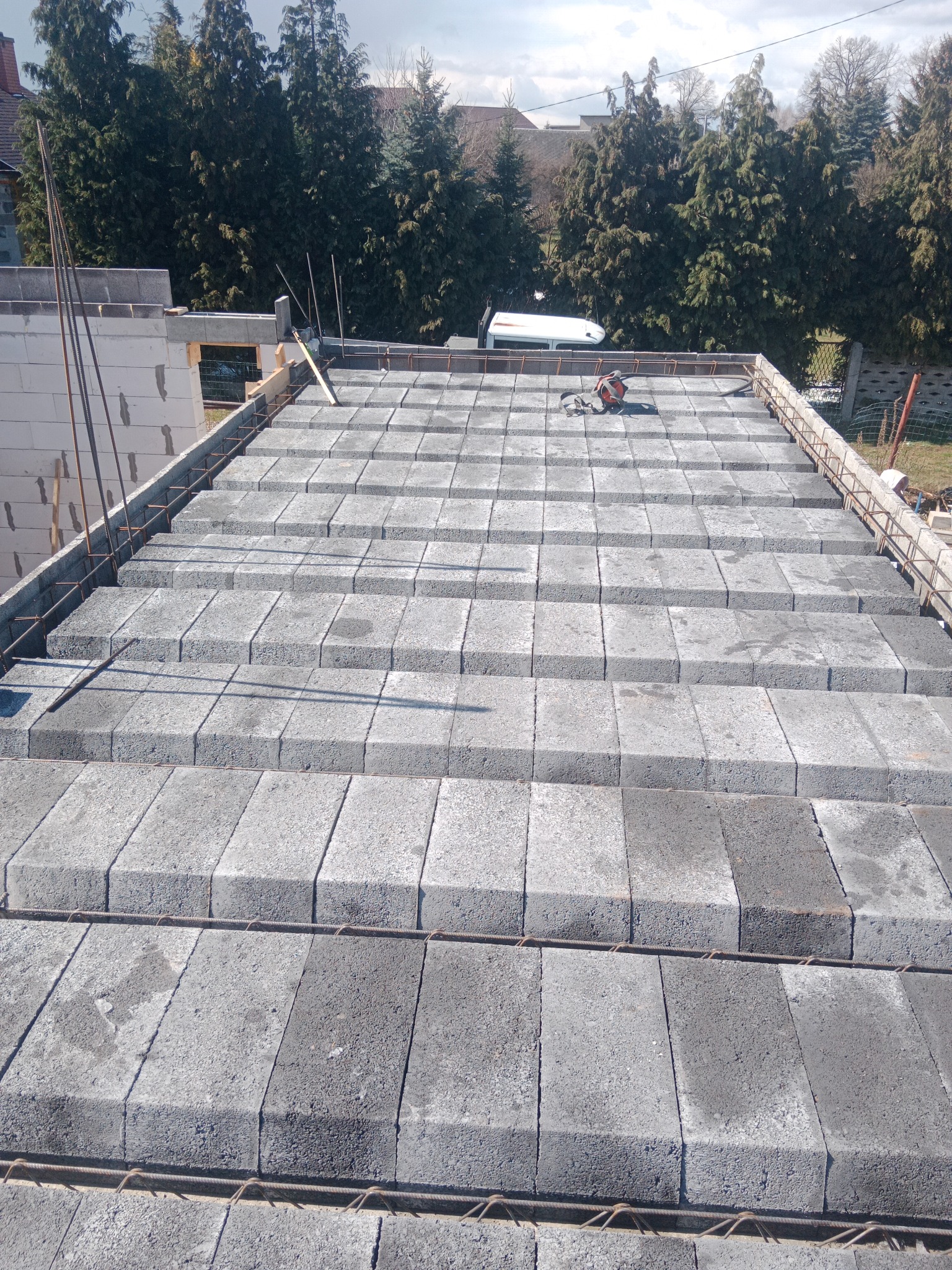 Wznoszenie stropu Teriva z betonowych pustaków i zbrojenia stalowego, widoczne elementy szalunku i narzędzia na budowie, w tle zabudowa jednorodzinna i drzewa.