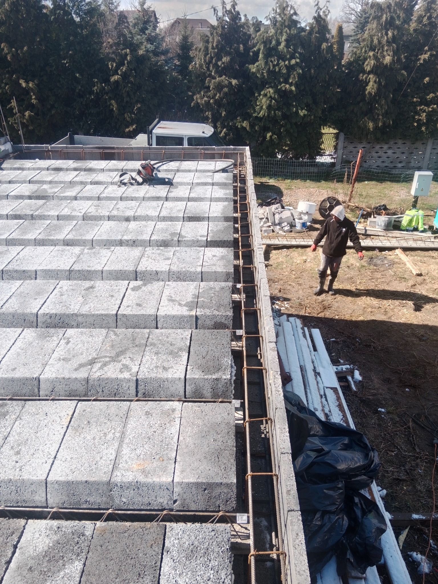 Wykonywanie stropu Teriva z betonowych pustaków i zbrojenia, widoczna osoba w roboczym ubraniu na placu budowy, w tle samochód dostawczy i wysokie drzewa.