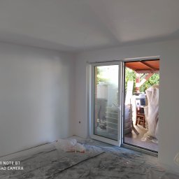 Rolsanders - Świeżo pomalowane na biało wnętrze pokoju z dużym oknem balkonowym; podłoga zabezpieczona folią malarską.