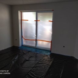 Rolsanders - Przygotowanie do prac w Lesznie: zabezpieczone folią okno i podłoga w pustym, świeżo wyszpachlowanym pomieszczeniu z widocznymi gniazdkami elektrycznymi.