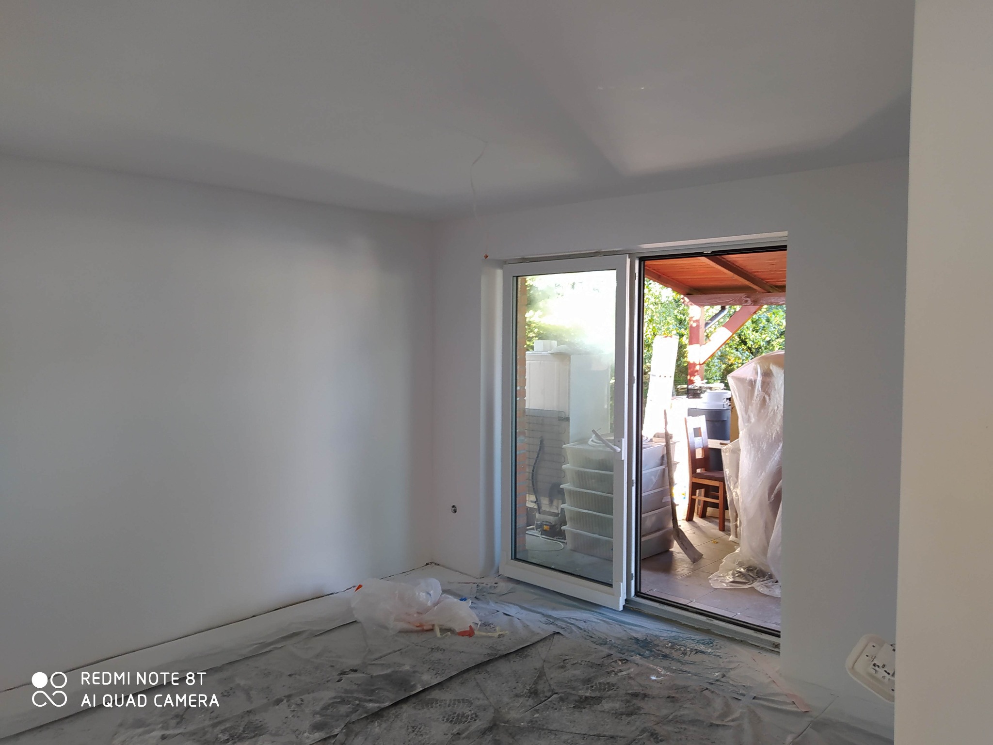 Świeżo pomalowane na biało wnętrze pokoju z dużym oknem balkonowym; podłoga zabezpieczona folią malarską.