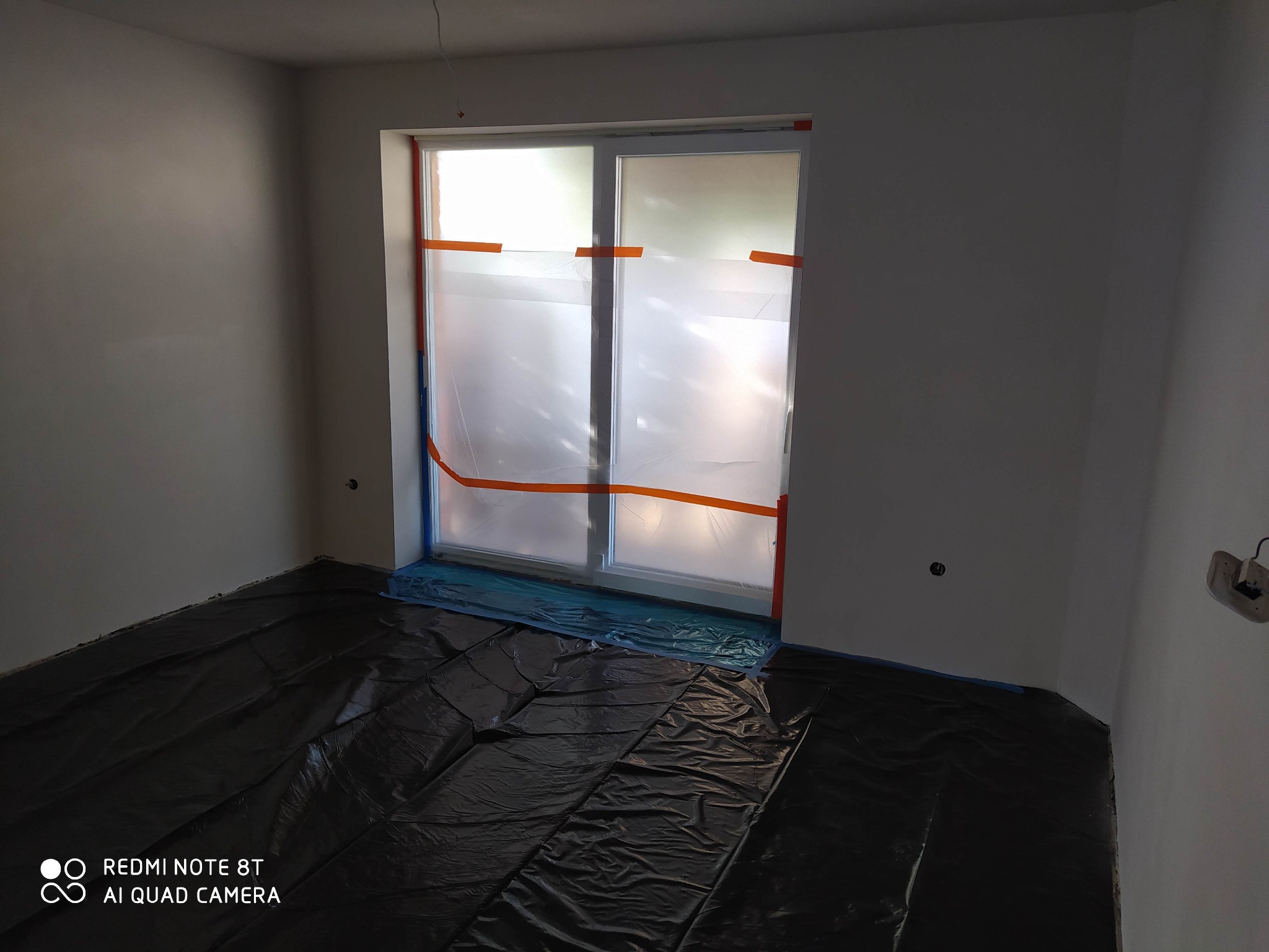 Przygotowanie do prac w Lesznie: zabezpieczone folią okno i podłoga w pustym, świeżo wyszpachlowanym pomieszczeniu z widocznymi gniazdkami elektrycznymi.