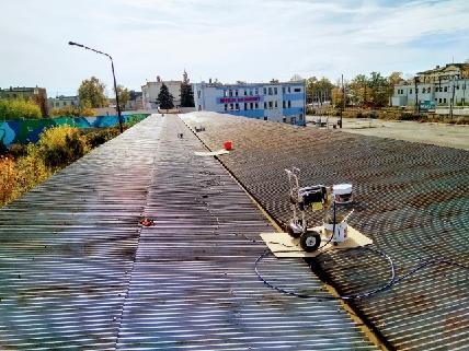 Agregat natryskowy na kartonie, przygotowany do pracy na falistym dachu budynku przemysłowego, widoczne ślady wcześniejszych prac i wiadro z farbą w tle.