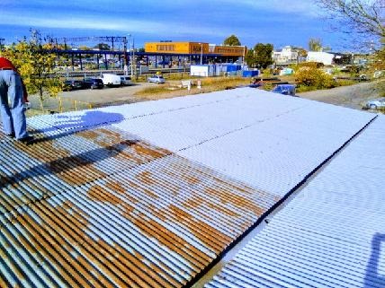 Pracownik w jeansach i czerwonej koszulce stoi na dachu z blachy falistej, częściowo pokrytym świeżą farbą, z widocznymi śladami rdzy na niepomalowanej powierzchni, w tle budynki przemysłowe...