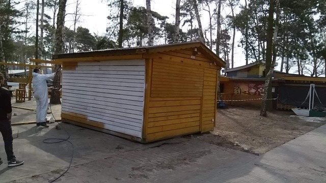 Pracownik w białym kombinezonie natryskowo nanosi farbę na drewniany kiosk, część ściany jest już biała, a część w kolorze drewna, otoczenie to park z drzewami.