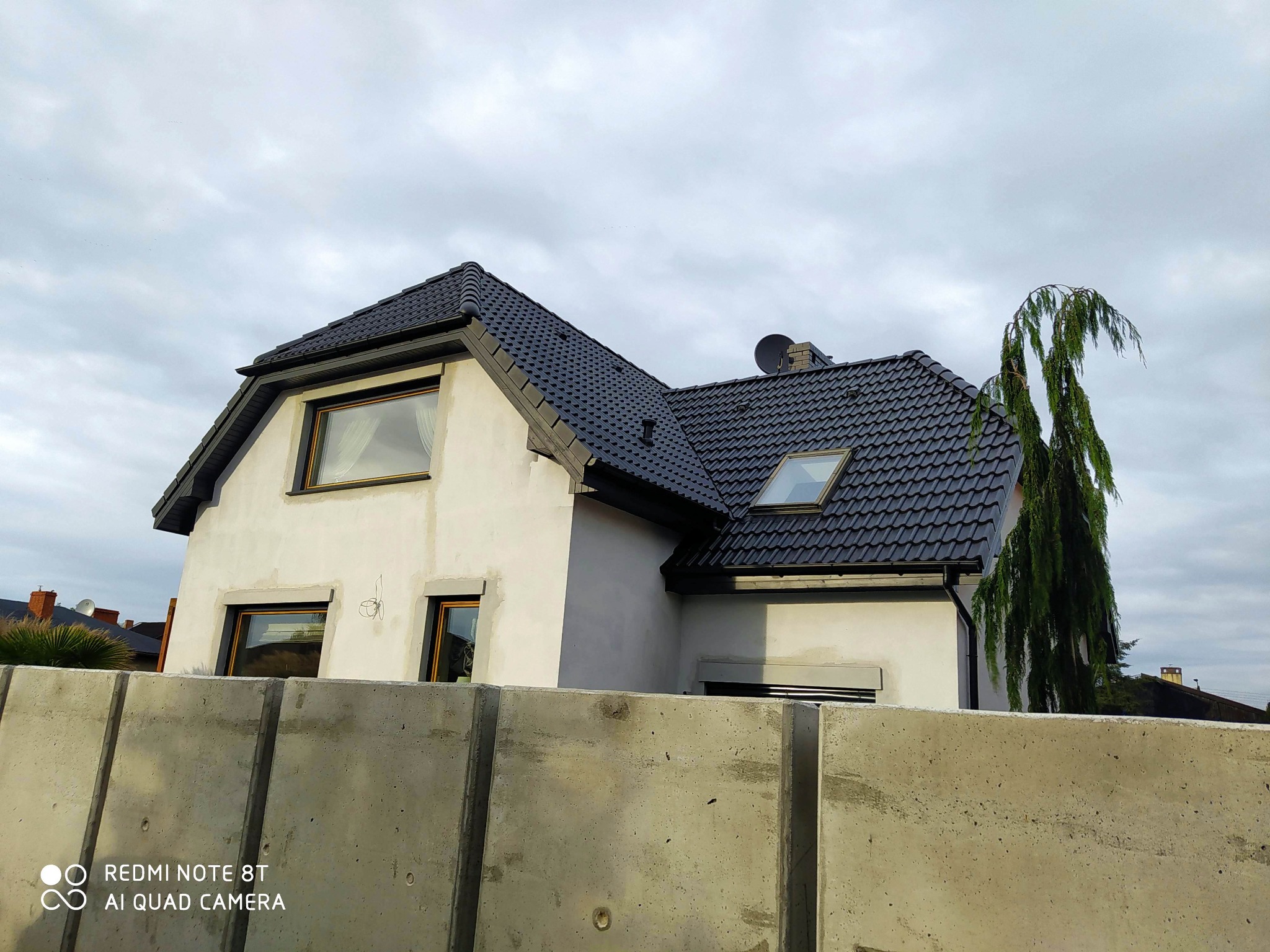 Surowa elewacja domu jednorodzinnego w trakcie malowania, widoczne fragmenty niepomalowane, czarny dach z dachówką, betonowe ogrodzenie na pierwszym planie.