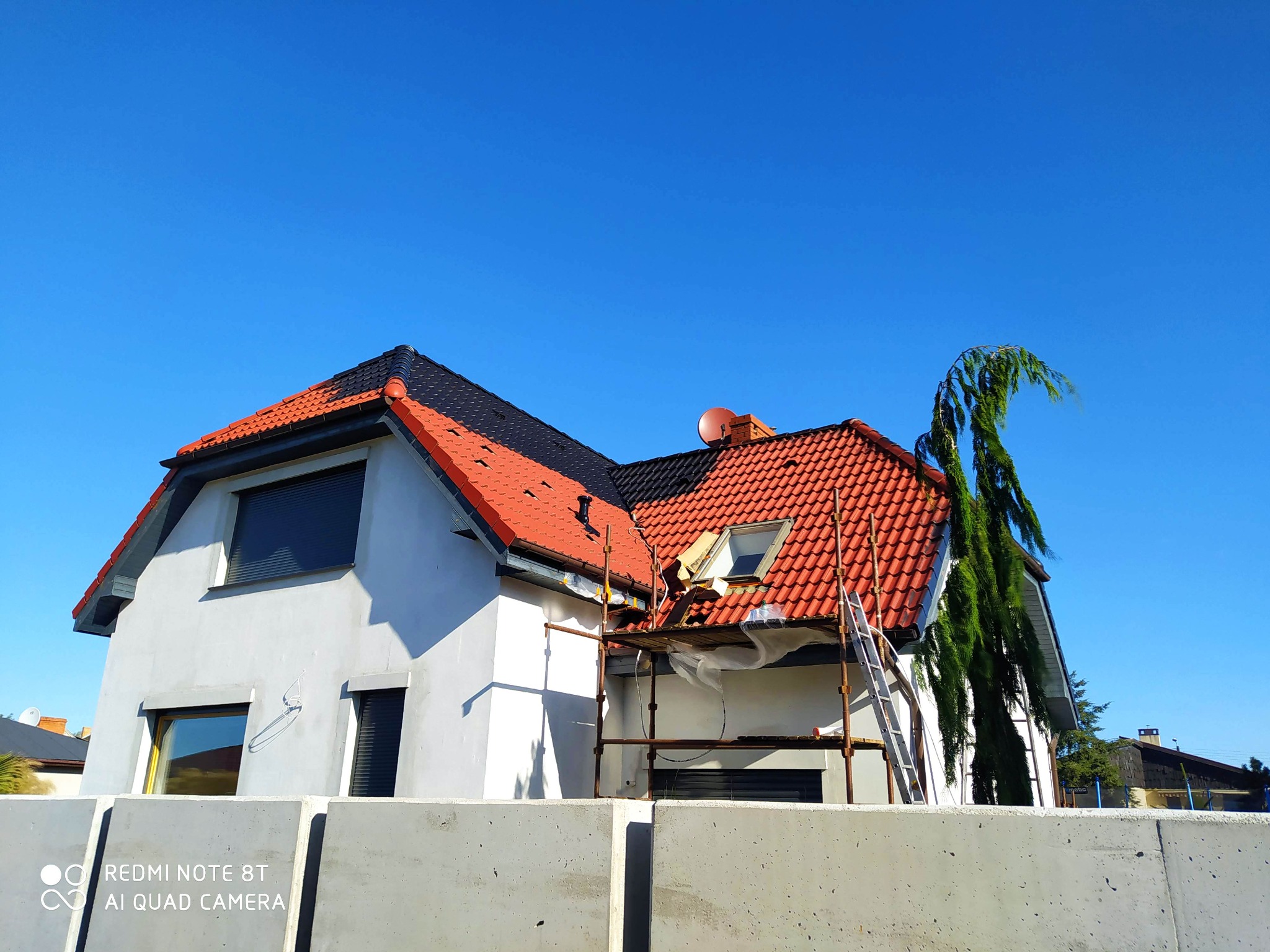 Dwukondygnacyjny dom jednorodzinny w trakcie prac na elewacji, widoczne rusztowanie, częściowo zakryte folią, oraz drabina oparta o ścianę; część dachu pokryta czerwoną dachówką, część czarną...