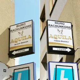 Szyld reklamowy biura rachunkowego 'Aquila' z logo ptaka, umieszczony na elewacji budynku, wskazujący kierunek na pierwsze piętro.