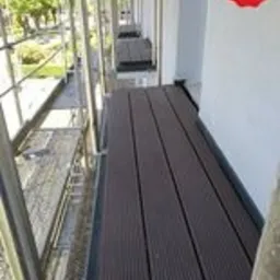 Ujęcie z rusztowania na elewacji budynku, widoczne brązowe deski kompozytowe ułożone na podeście roboczym, w tle zieleń drzew i chodnik.