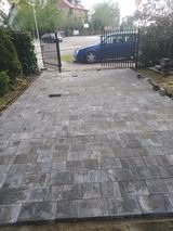 Ułożona kostka brukowa na podjeździe, widoczne ślady po deszczu, w tle brama wjazdowa i zaparkowany granatowy samochód.