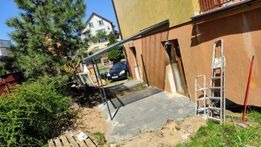 Elewacja domu jednorodzinnego w trakcie prac wykończeniowych, widoczne ocieplenie, przygotowane do nałożenia tynku, drabina oparta o ścianę, teren wokół budynku częściowo rozkopany.