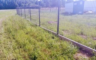 Długi, ciemny metalowy płot panelowy z podmurówką betonową, biegnący wzdłuż zarośniętego trawnika w kierunku oddalonych drzew i budynku gospodarczego w tle.