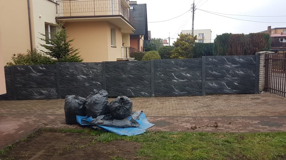 Nowo postawiony, ciemnoszary płot betonowy o fakturze imitującej kamień, widoczny z perspektywy trawnika z workami odpadów budowlanych i rozłożoną plandeką.