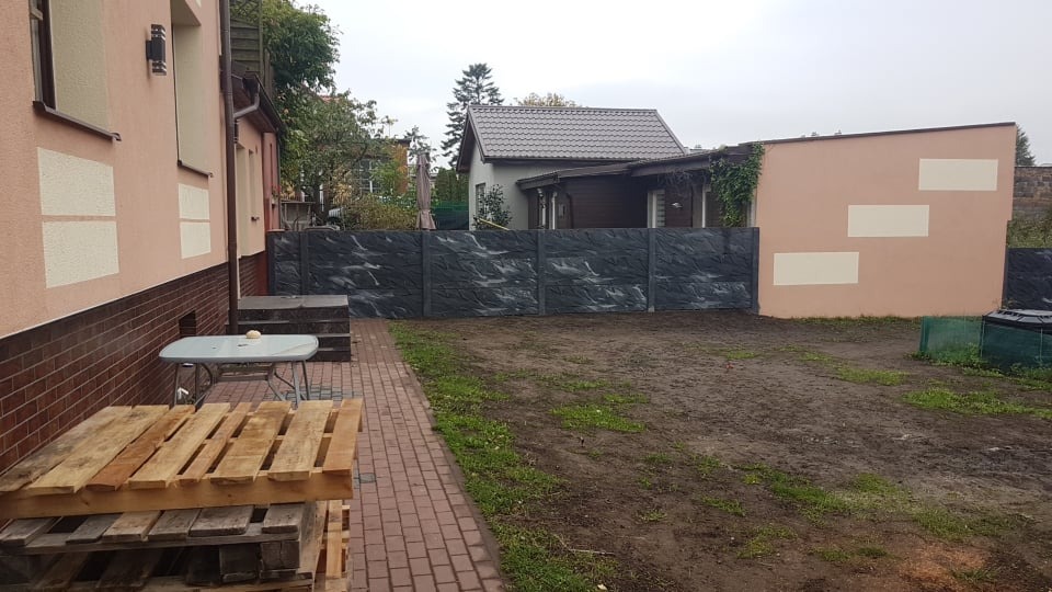Nowoczesny, szary płot z imitacją kamienia, oddzielający posesję w Pucku. Widoczny fragment domu z elewacją w odcieniach beżu i brązu oraz paleta drewniana na pierwszym planie.
