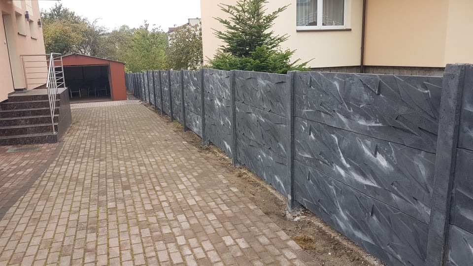 Długa, szara, betonowa ściana z wyraźną fakturą, oddzielająca posesję od chodnika z kostki brukowej; w tle budynek mieszkalny i garaż.
