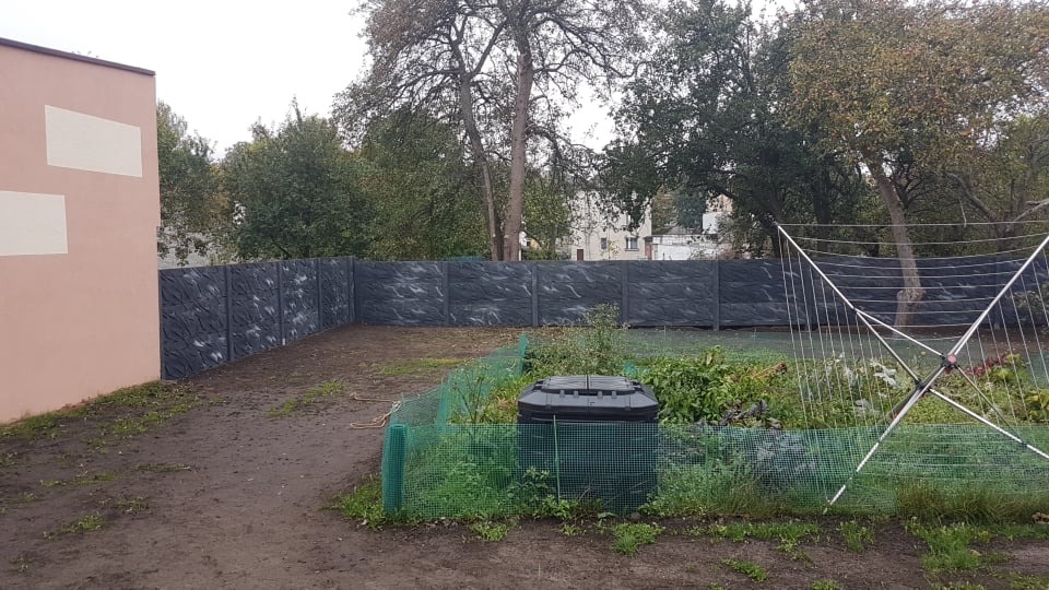Nowoczesne, grafitowe ogrodzenie panelowe z wyraźną fakturą kamienia, otaczające ogród z kompostownikiem i rozłożoną suszarką na pranie, przylegające do budynku z jasnymi wstawkami elewacyjnymi.
