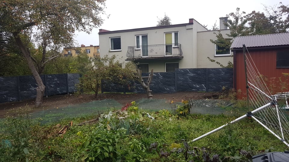 Dwupiętrowy dom z balkonem, otoczony nowym, szarym ogrodzeniem imitującym kamień, widoczny zarośnięty ogród z rozłożoną suszarką na pranie i małą czerwoną szopą.