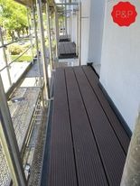 Ujęcie z rusztowania na elewacji budynku, widoczne brązowe deski kompozytowe ułożone na podeście roboczym, w tle zieleń drzew i chodnik.