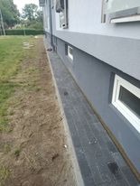 Wykończona elewacja domu jednorodzinnego z szarą podmurówką i jasnoszarą ścianą, widoczne małe okienka piwniczne, trawnik w tle.