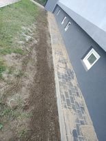 Ciemnoszara elewacja budynku z widocznymi białymi oknami, przylegająca do nierównego terenu z częściowo ułożoną kostką brukową i pozostałościami piasku lub zaprawy.