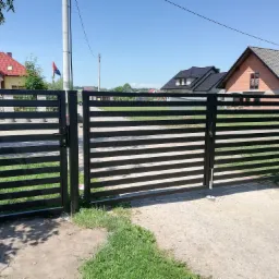 Nowoczesna, pozioma brama wjazdowa z czarnych profili stalowych na tle domów jednorodzinnych i błękitnego nieba.
