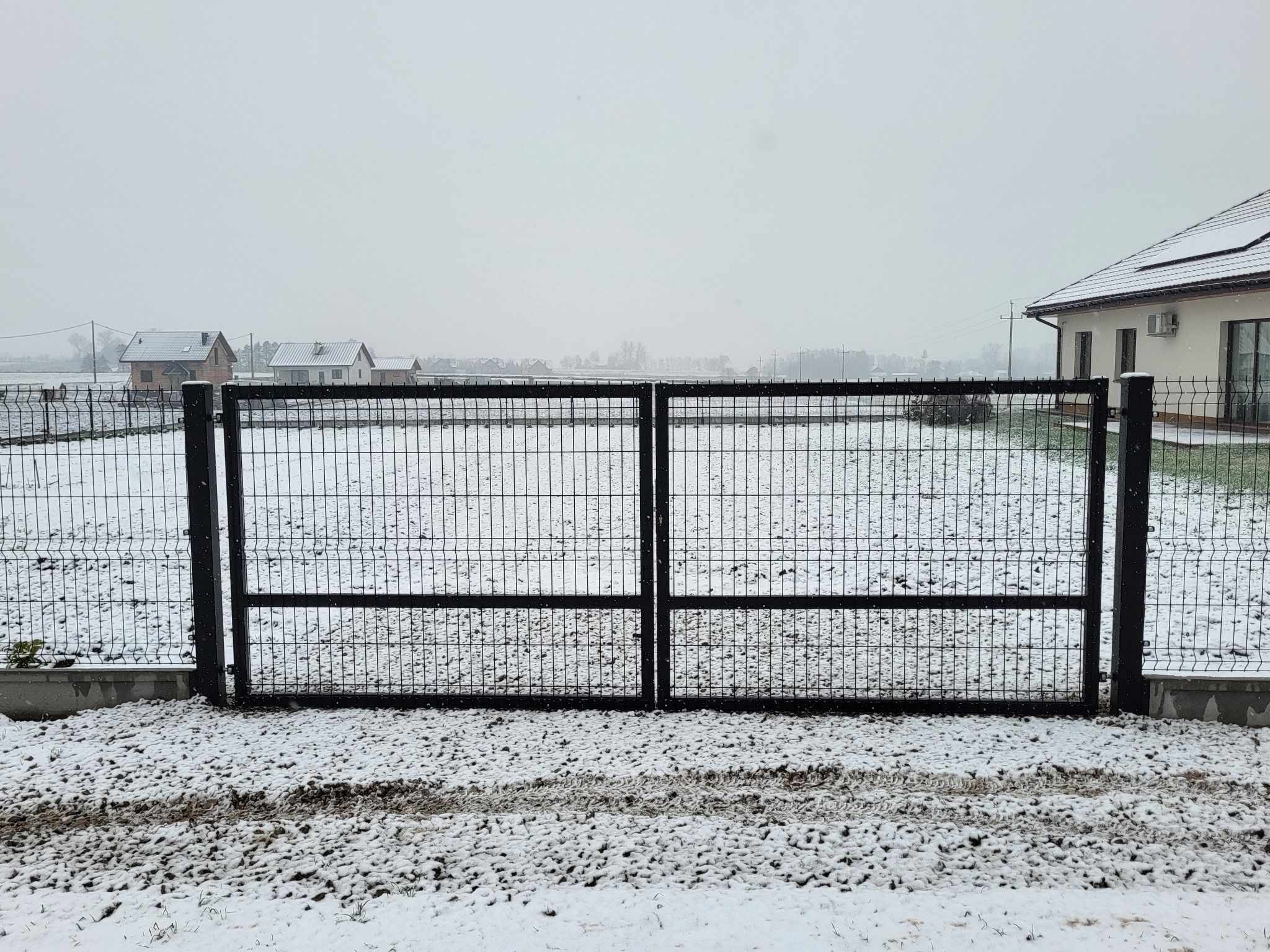 Dwuskrzydłowa, metalowa brama z siatki, pomalowana na czarno, w zimowej scenerii. Świeżo spadły śnieg pokrywa ziemię i częściowo przylega do siatki bramy. W tle widoczne domy jednorodzinne.