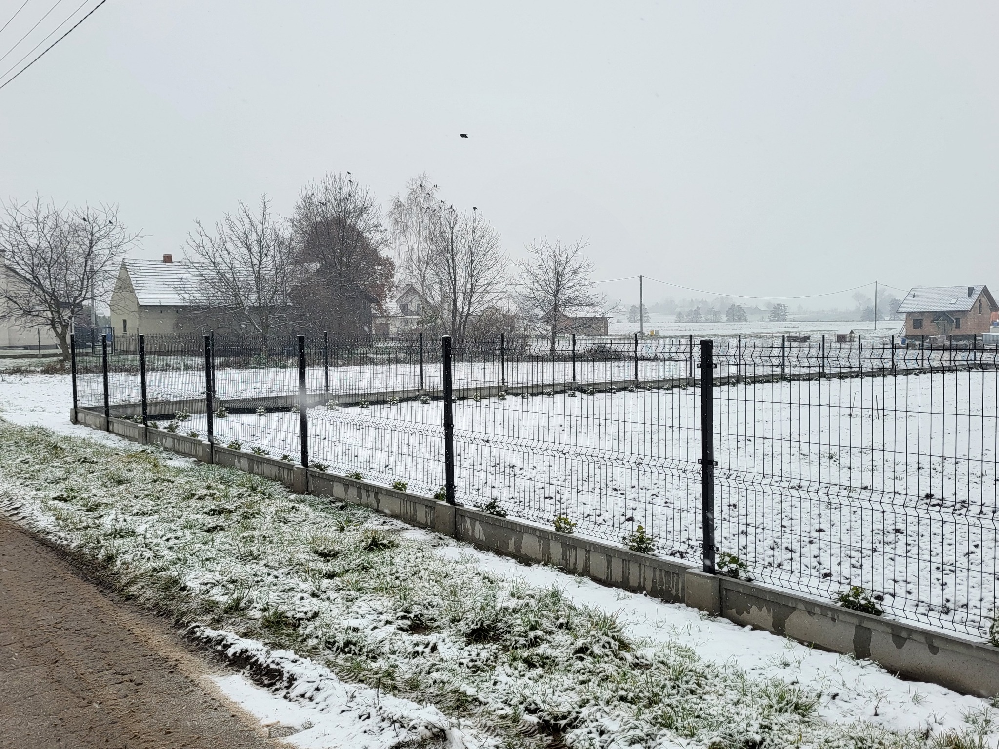 Nowo postawione, czarne ogrodzenie panelowe na betonowej podmurówce, częściowo pokryte śniegiem na tle zaśnieżonego pola i zabudowań w pochmurny dzień.