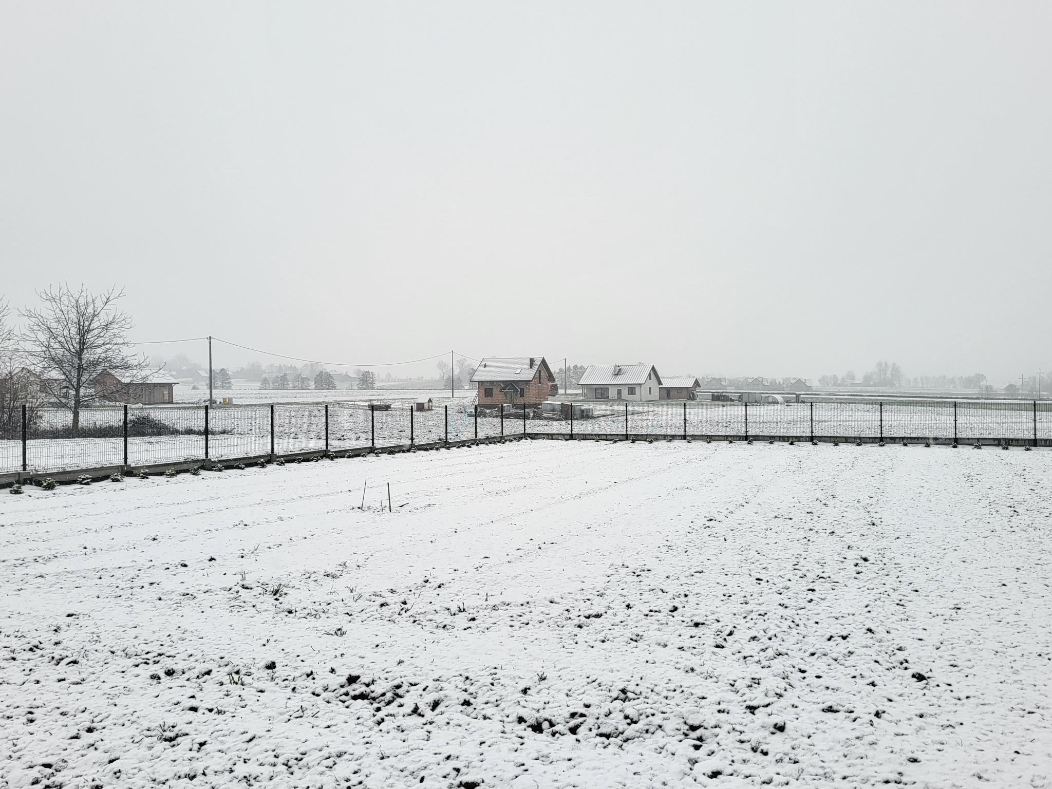 Metalowe ogrodzenie panelowe otaczające zaśnieżone pole uprawne z widokiem na domy w oddali, pochmurny zimowy dzień.