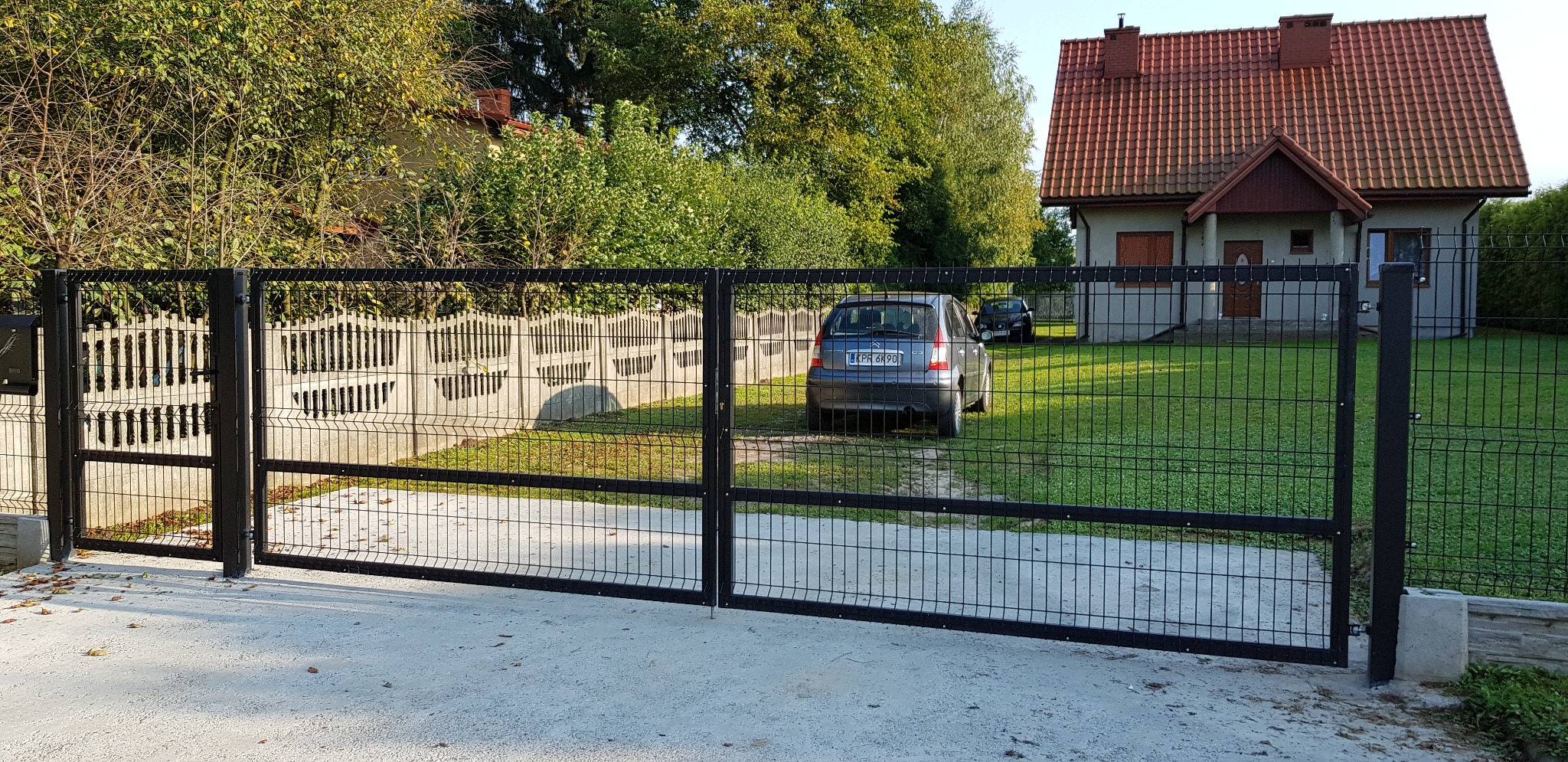 Nowoczesna, czarna, metalowa brama dwuskrzydłowa z siatki panelowej przed domem z czerwoną dachówką; w tle zaparkowane dwa samochody.