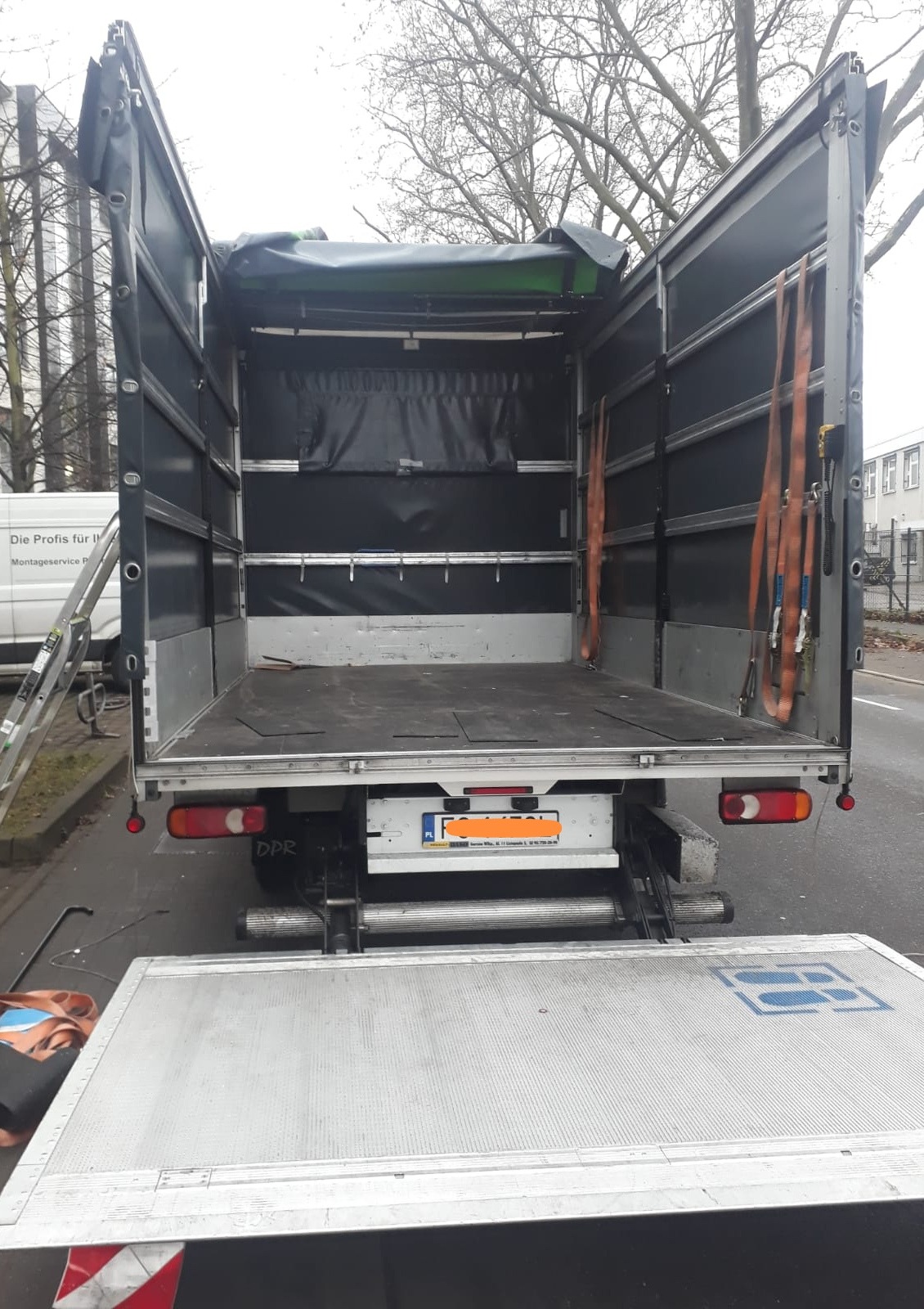 Otwarta pusta przestrzeń ładunkowa samochodu dostawczego z windą załadowczą, widoczne pasy mocujące po bokach, drabina oparta o bok pojazdu.