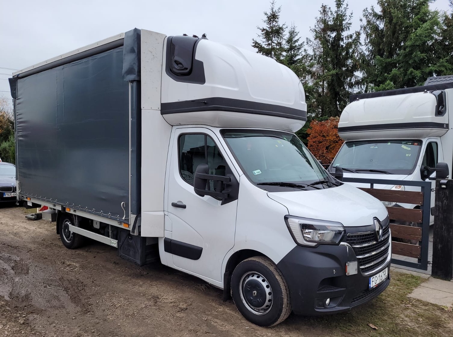 Biały bus Renault Master z plandeką na błotnistym podłożu, widoczny drugi podobny bus w tle, na tablicy rejestracyjnej widnieje FG 1679L.