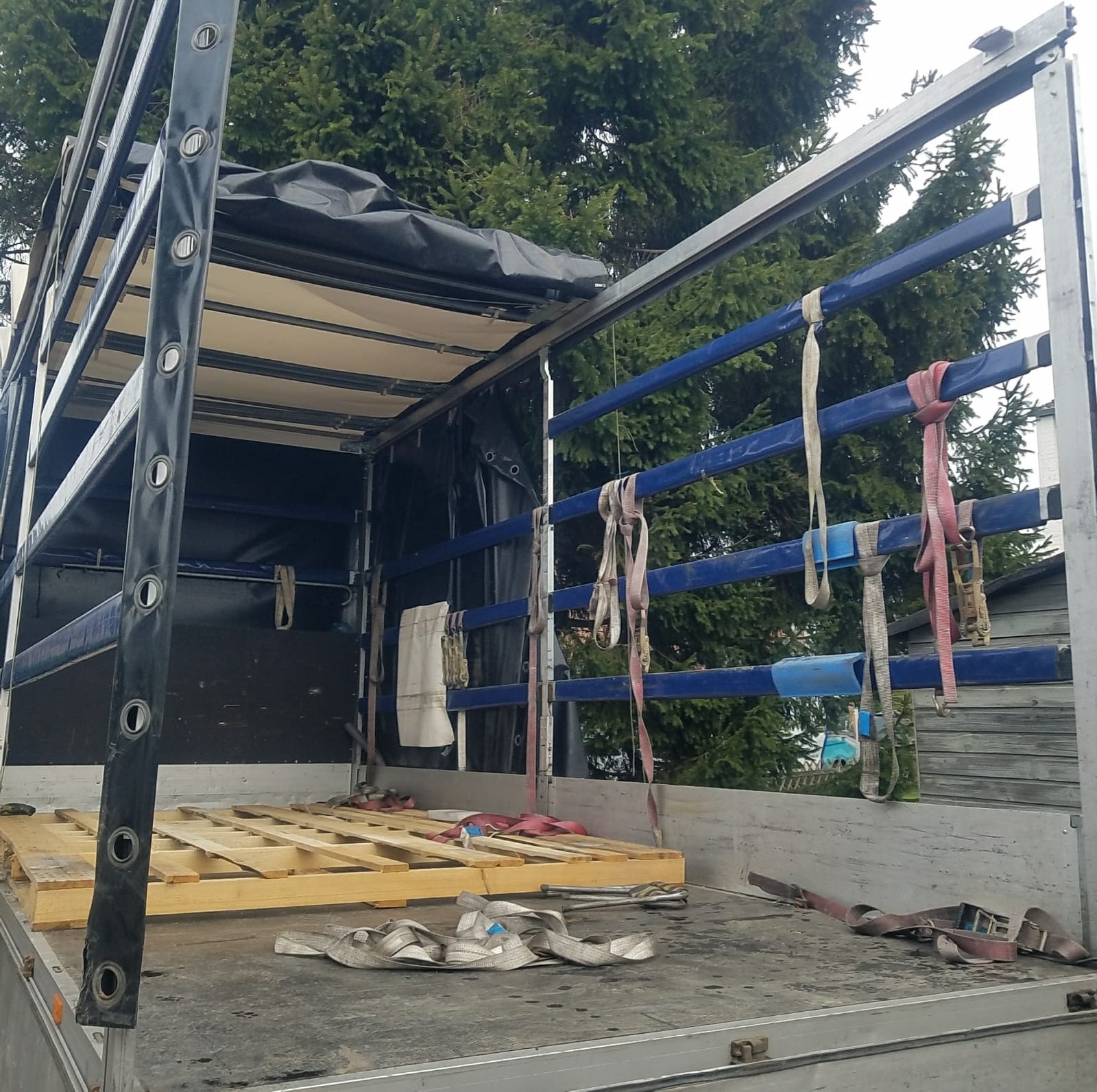 Wnętrze skrzyni ładunkowej samochodu dostawczego z drewnianą paletą na podłodze i pasami mocującymi zwisającymi z boku, widok na zewnątrz na zieleń drzew.