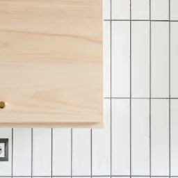 Jasne, drewniane szafki kuchenne z okrągłymi, złotymi uchwytami, zamontowane nad potrójnym gniazdkiem elektrycznym w czarnej ramce, na tle białych, wąskich płytek ułożonych pionowo z ciemną fugą.