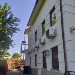 Thnck Group - Biała elewacja budynku z zamontowanymi klimatyzatorami, widoczne okna, rynna, rusztowanie, kosz na śmieci i mur z cegieł.