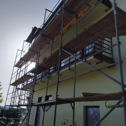 Thnck Group - Dom w trakcie remontu elewacji, otoczony rusztowaniem, z widocznym drzewem w tle.