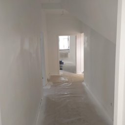 Thnck Group - Korytarz w trakcie malowania na biało, podłoga zabezpieczona folią, widoczne kable wystające z sufitu, pokój w głębi z grzejnikiem pod oknem.