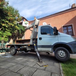 Thnck Group - Podnośnik koszowy na pojeździe ustawiony na chodniku, z pracownikiem w koszu na tle budynku mieszkalnego i drzew, sugeruje prace na wysokościach.