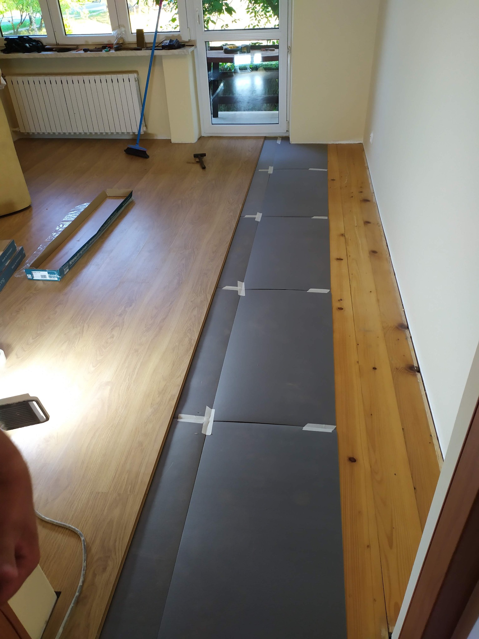Fragment pokoju w trakcie układania paneli podłogowych: część podłogi wyłożona szarą pianką z taśmą klejącą, obok ułożone panele, w tle widoczny otwarty karton z panelami i narzędzia.