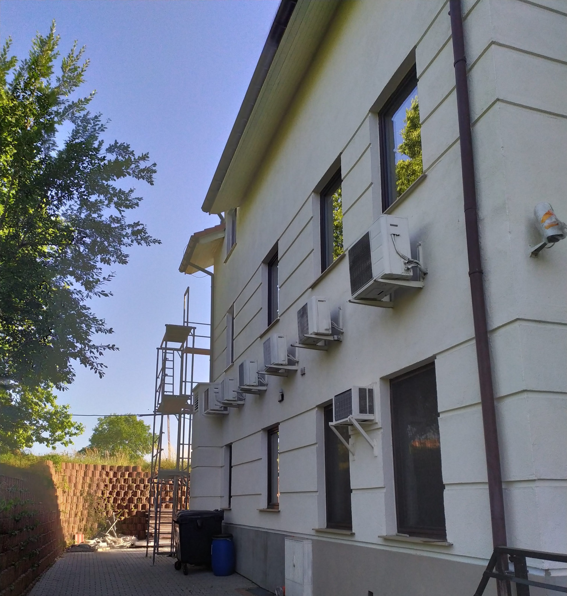 Biała elewacja budynku z zamontowanymi klimatyzatorami, widoczne okna, rynna, rusztowanie, kosz na śmieci i mur z cegieł.