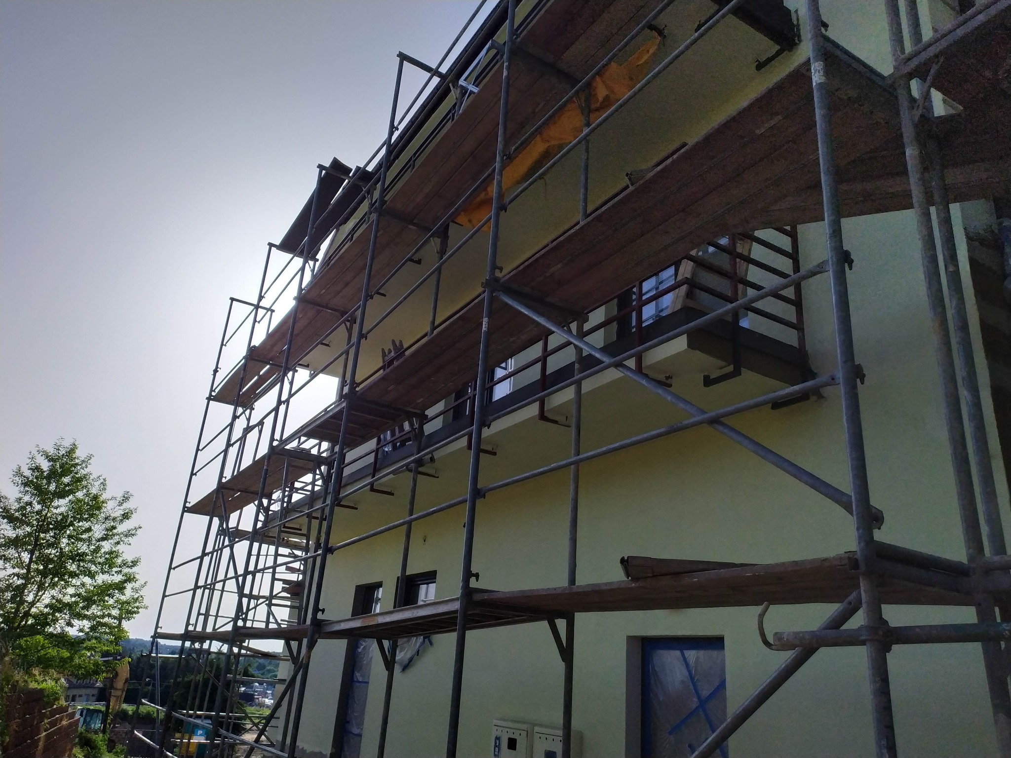 Dom w trakcie remontu elewacji, otoczony rusztowaniem, z widocznym drzewem w tle.