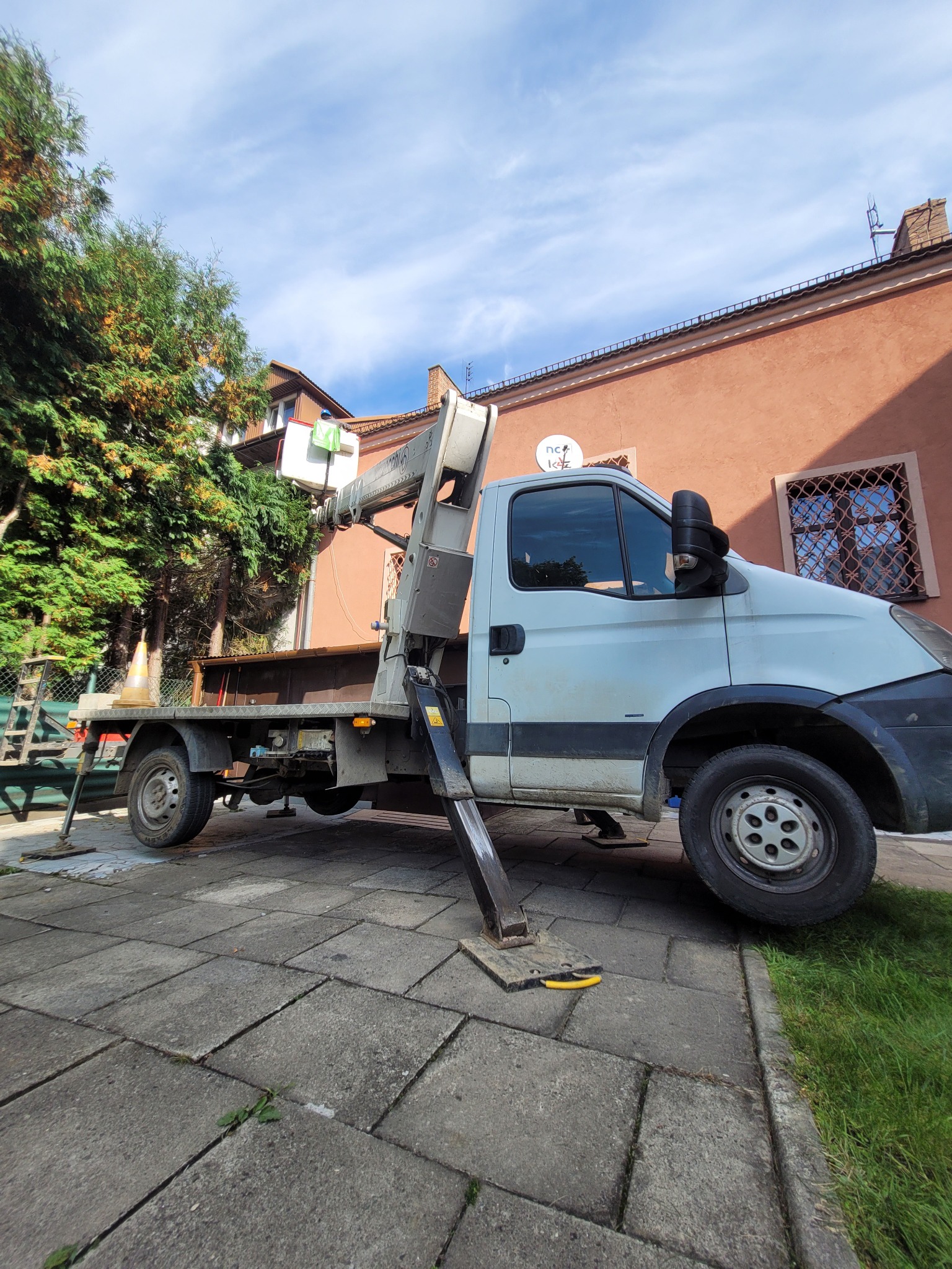 Podnośnik koszowy na pojeździe ustawiony na chodniku, z pracownikiem w koszu na tle budynku mieszkalnego i drzew, sugeruje prace na wysokościach.