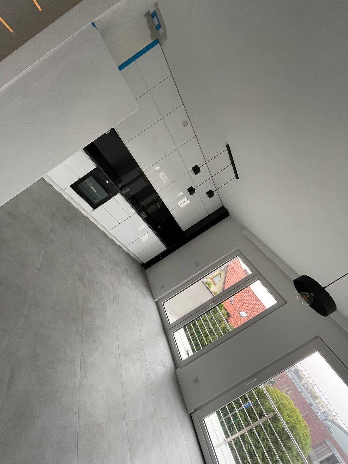 Minimalistyczna kuchnia z białymi szafkami i czarnymi akcentami w Krakowie, widok z dołu ukazuje szare płytki podłogowe i okna z widokiem na zieleń.