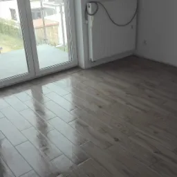 Błyszcząca, świeżo ułożona podłoga z płytek imitujących drewno w pustym pokoju z grzejnikiem i wyjściem na balkon.