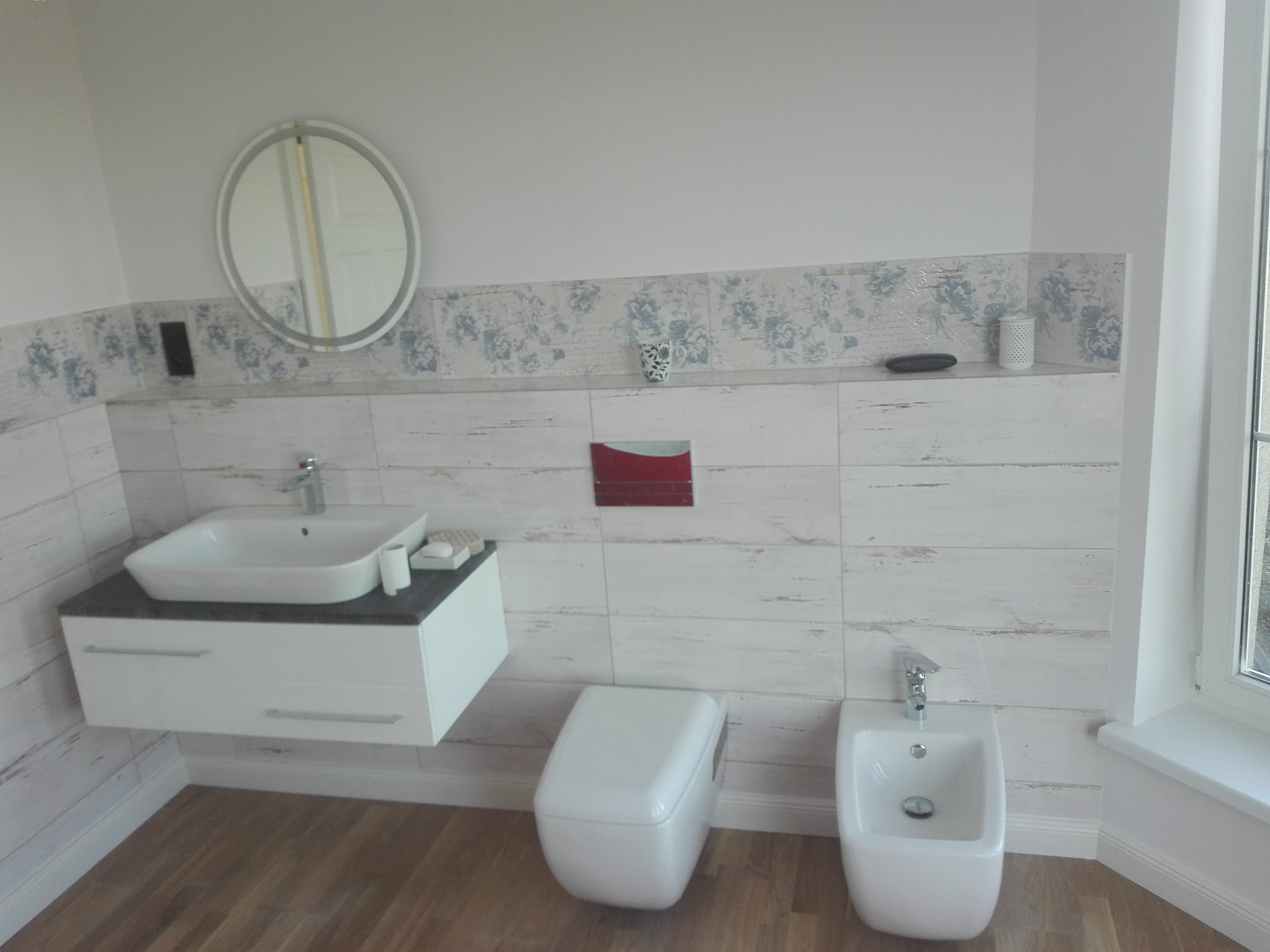 Nowoczesna łazienka z drewnopodobnymi płytkami, wiszącą szafką z umywalką, podwieszaną toaletą i bidetem, ozdobiona lustrem z oświetleniem i dekoracyjnym pasem z motywem kwiatowym.