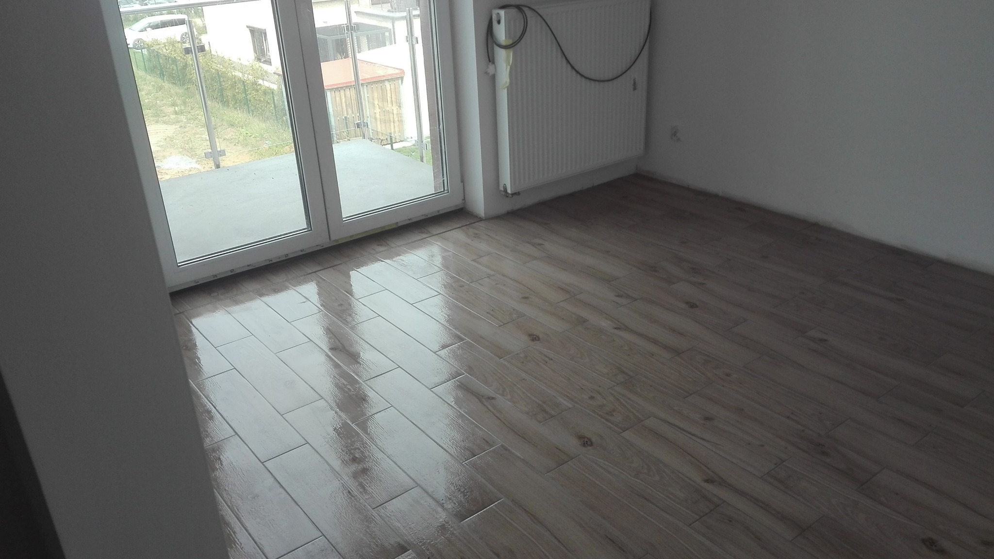 Błyszcząca, świeżo ułożona podłoga z płytek imitujących drewno w pustym pokoju z grzejnikiem i wyjściem na balkon.