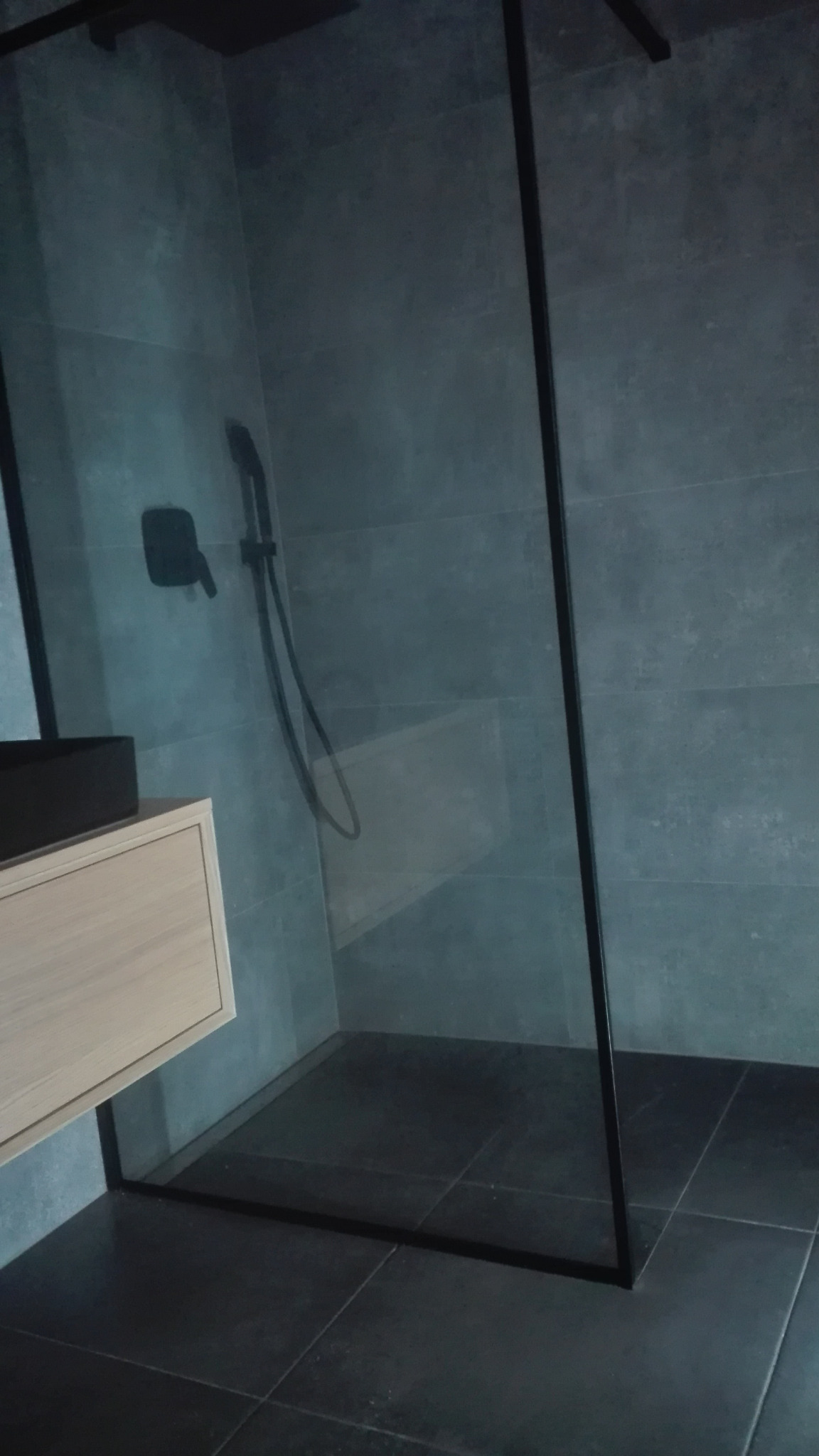 Nowoczesna łazienka z kabiną prysznicową typu walk-in z czarnymi profilami, szara glazura i drewniana szafka pod umywalką.