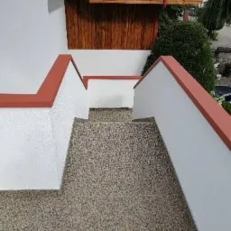 Zewnętrzne schody wejściowe do domu z wykończeniem z żywicy epoksydowej w odcieniach szarości, białe ściany z czerwoną lamówką, drewniane zadaszenie nad wejściem.