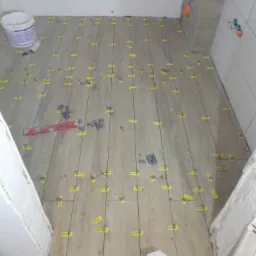 Układanie płytek podłogowych imitujących drewno w łazience, z użyciem żółtych klipsów poziomujących i poziomicy, widoczny biały pojemnik po kleju.