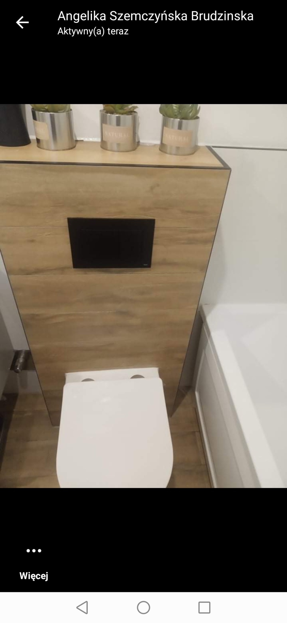 Wisząca toaleta z zabudową z płyty drewnopodobnej, czarny panel sterujący, trzy srebrne doniczki z roślinami na górze, biała wanna po prawej stronie.