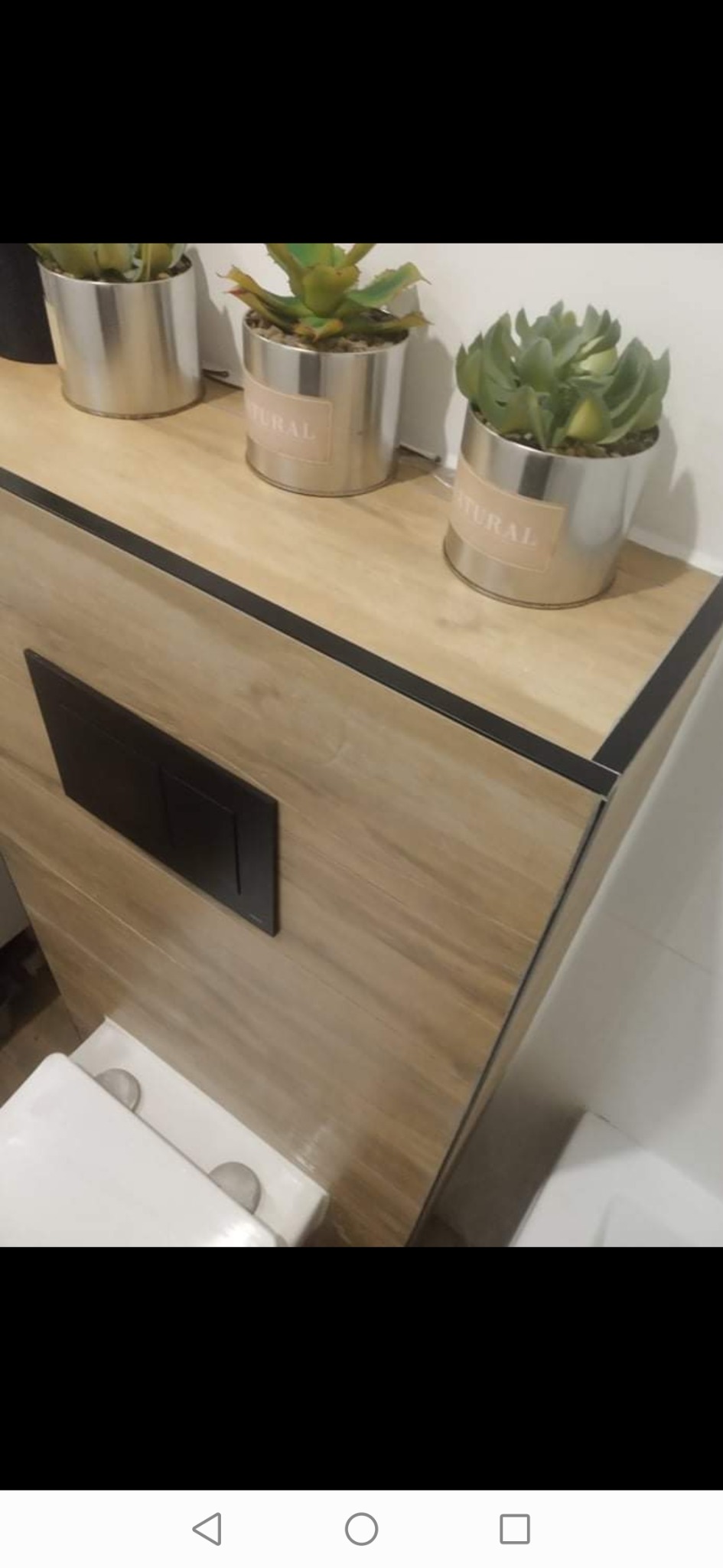 Nowoczesna toaleta z zabudowaną spłuczką i blatem wykończonym płytkami imitującymi drewno, na którym stoją trzy srebrne doniczki z roślinami.